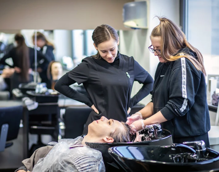 Two hairstyling students washing a client's hair at a salon sink. Two hairstyling students washing a client's hair at a salon sink.